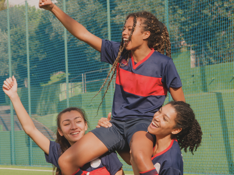 Trois jeunes filles célèbrent leur victoire au foot, l'une est portée sur les épaules des deux autres.