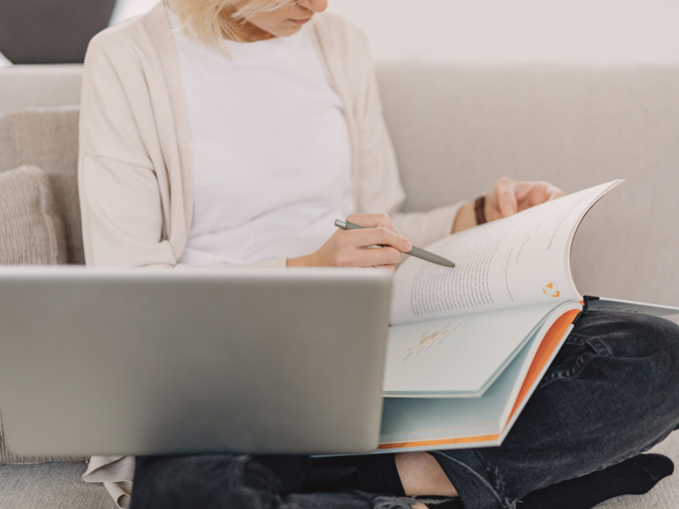 Une femme révise sur son canapé à l'aide d'un livre et de son ordinateur portable.