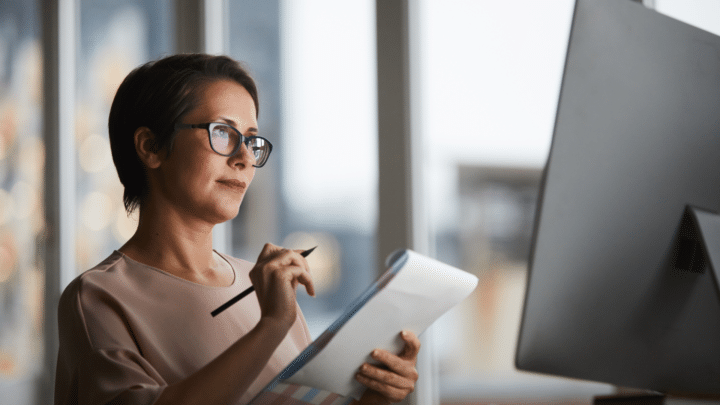 Une femme regarde son écran d’ordinateur tout en prenant des notes sur un bloc.