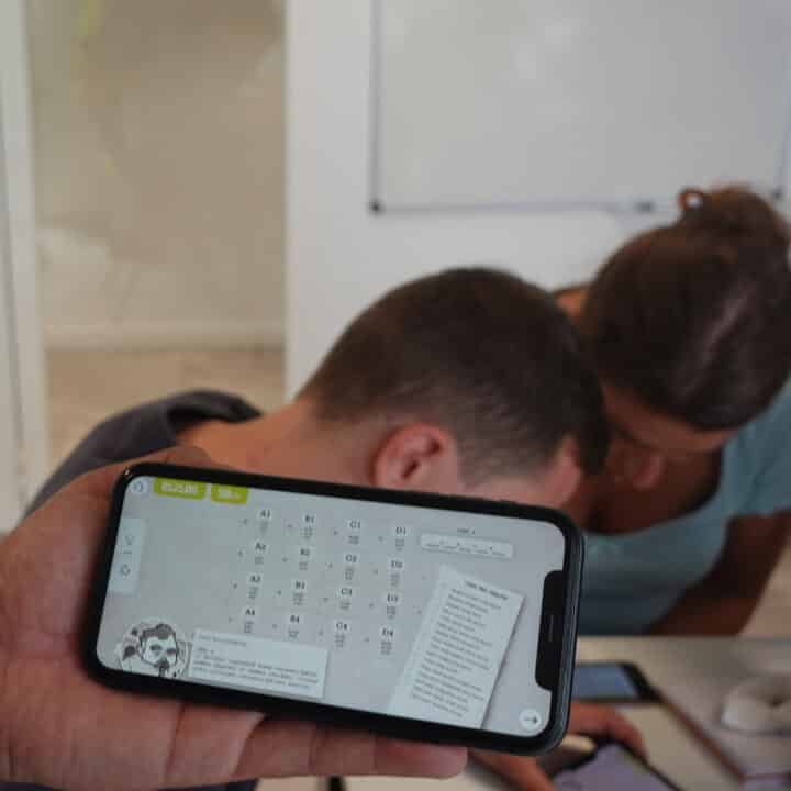Une personne tient un smartphone affichant une application éducative, tandis que deux jeunes adultes se concentrent ensemble sur un autre téléphone en arrière-plan.