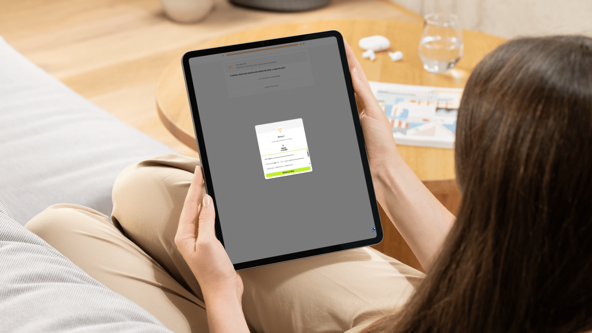 Une jeune femme utilise une tablette pour suivre un exercice de français en ligne, confortablement installée chez elle.