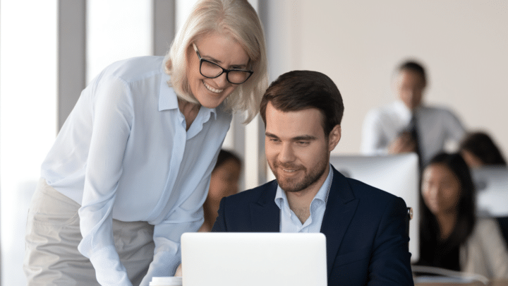 Une formatrice souriante accompagne un homme en costume sur son ordinateur dans un environnement professionnel, en salle de formation.