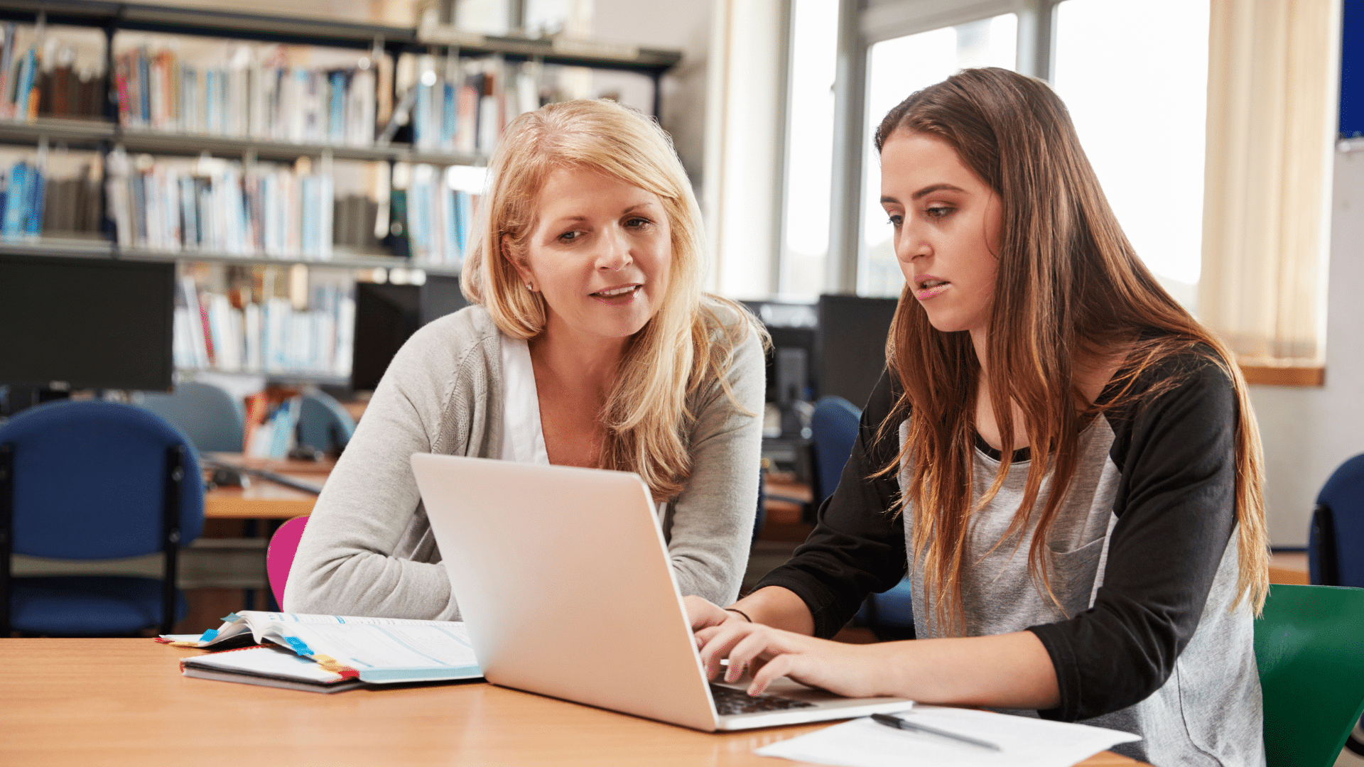 Une enseignante accompagne une élève dans son travail sur ordinateur, dans une salle équipée de livres et d’ordinateurs.