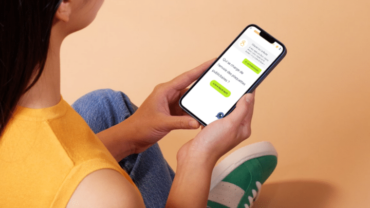 Une jeune femme consulte un exercice d’orthographe Projet Voltaire sur son smartphone, assise contre un mur saumon, jambes repliées.